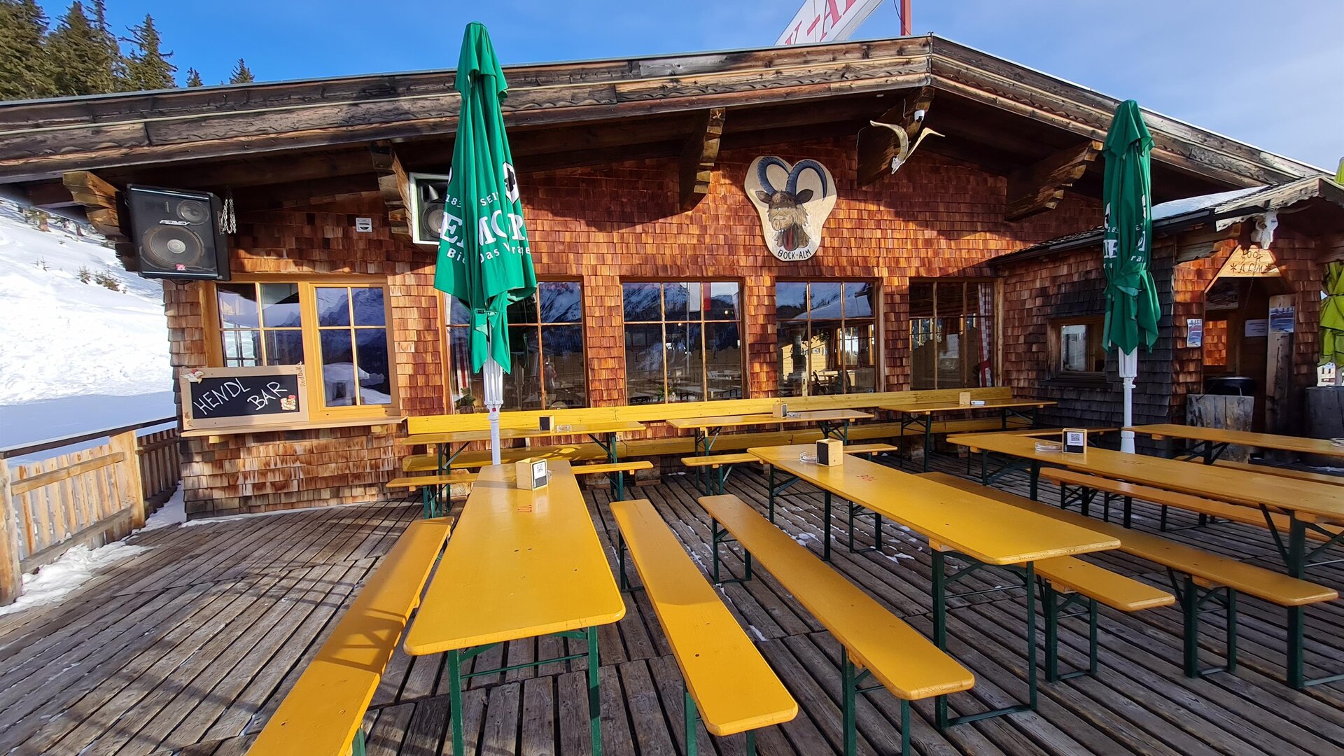 Eine rustikale Berghütte mit einer Holzterrasse und gelben Bänken. Es gibt grüne Sonnenschirme und verschneite Landschaft im Hintergrund.