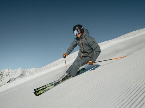 Ein Skifahrer fährt über die frisch präparierte Piste. Der Himmel ist klar und die Berge sind im Hintergrund sichtbar.