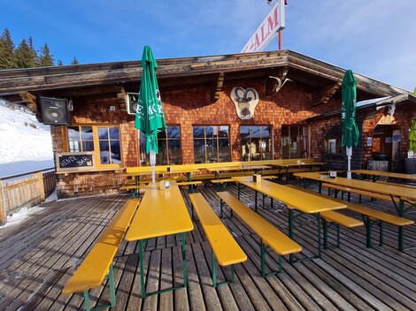 Eine gemütliche Berghütte mit Holzverkleidung und mehreren langen Bänken. Die Terrasse ist mit grünen Sonnenschirmen ausgestattet und der Schnee ist sichtbar im Hintergrund.