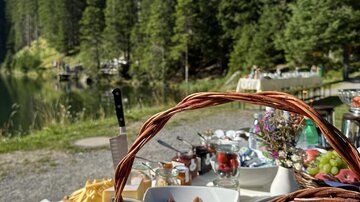 Ein schön gedeckter Tisch in der Natur mit frischen Croissants, Käse und Obst. Im Hintergrund sind hohe Bäume und ein klarer blauer Himmel zu sehen.