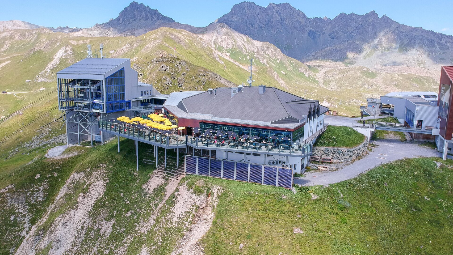 A modern mountain restaurant with a large terrace, surrounded by green meadows and mountains. The clear sky offers a dreamlike view of the alpine landscape.