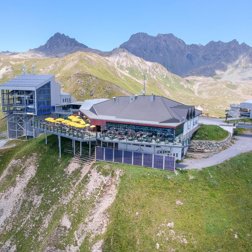 A modern mountain restaurant with a large terrace, surrounded by green meadows and mountains. The clear sky offers a dreamlike view of the alpine landscape.