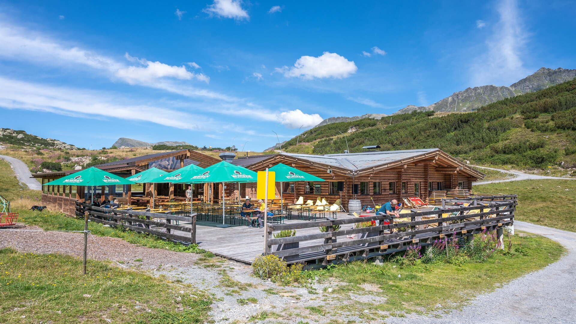 Ein gemütliches Holzhaus mit einer Terrasse und grünen Sonnenschirmen. Die Umgebung ist bergig und grün unter einem blauen Himmel.