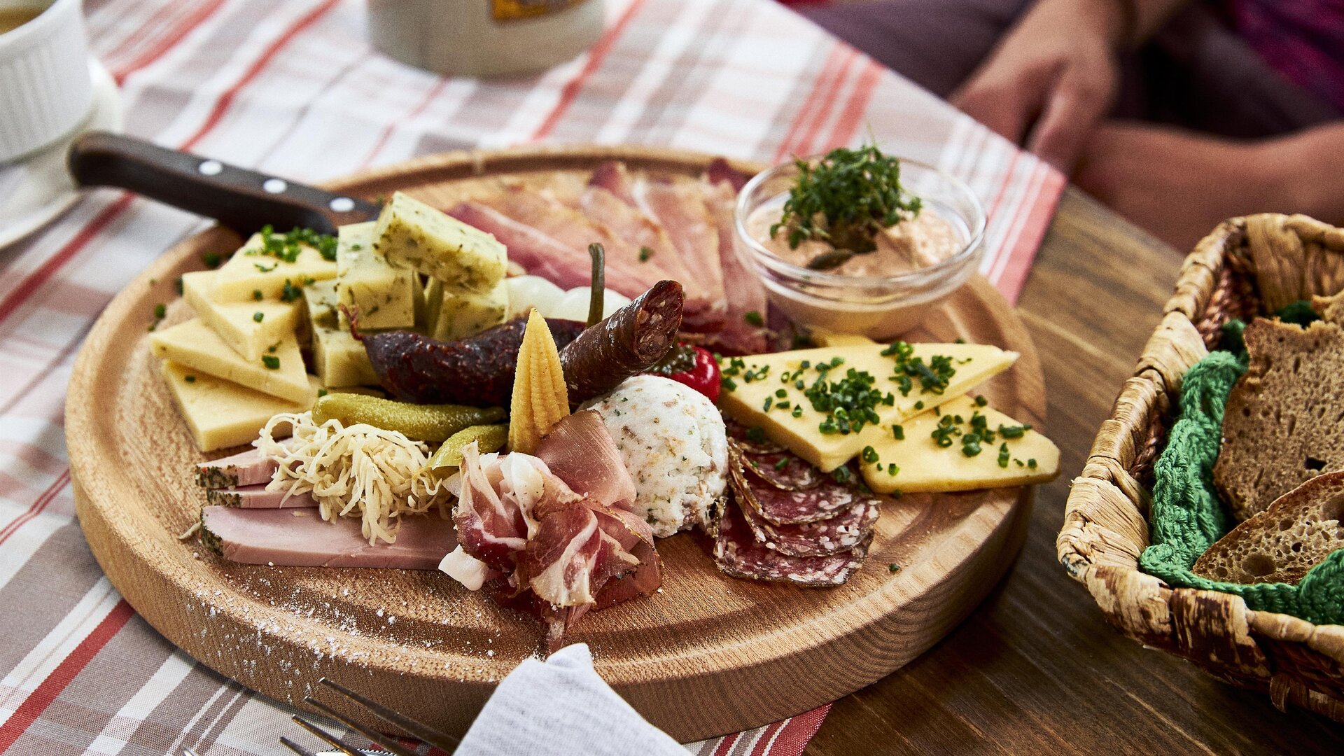 Eine rustikale Käse- und Wurstplatte auf einem Holzbrett. Dazu gibt es eingelegtes Gemüse und einen Dip im kleinen Schälchen.