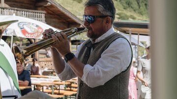 A man plays trumpet in traditional attire. In the background, tables and benches as well as green mountains can be seen.