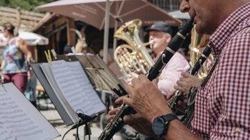 A band is playing outdoors under parasols. The man at the front is playing the clarinet and is wearing a checkered blouse.