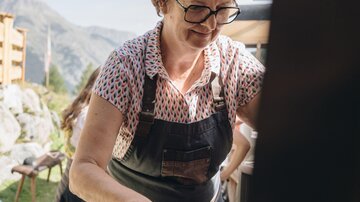 A woman in an apron is working intently at a table outdoors. In the background, mountains and a clear sky are visible.