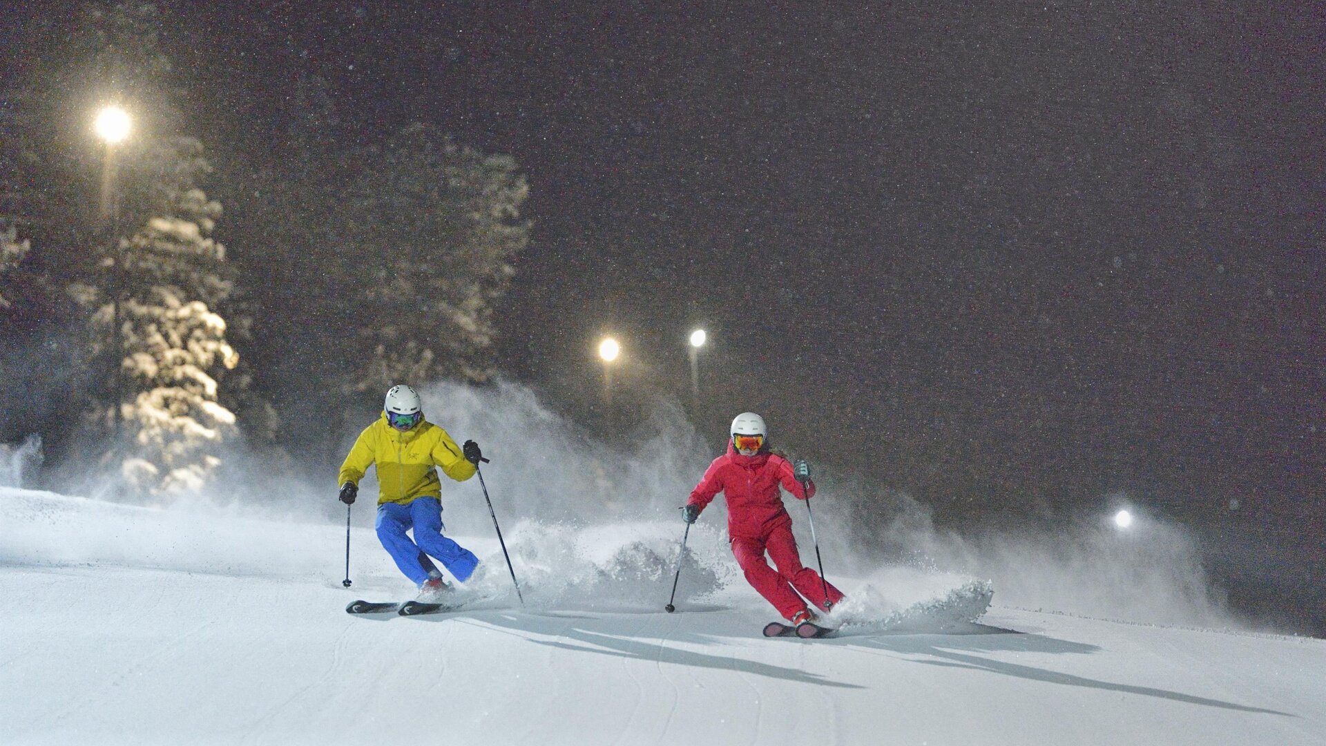 Zwei Skifahrer fahren nachts auf einer schneebedeckten Piste. Die Umgebung ist beleuchtet und erzeugt eine winterliche Atmosphäre.