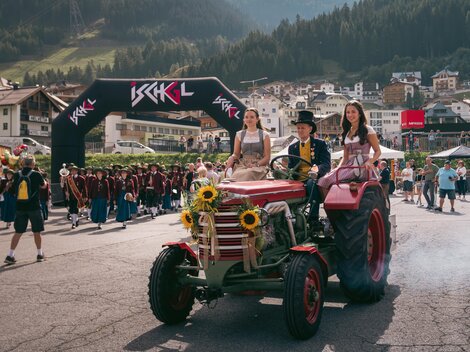 Eine lebhafte Veranstaltung mit einem Traktor, der von zwei Frauen in traditionellen Trachten begleitet wird. Im Hintergrund sind jubelnde Menschen und eine malerische Berglandschaft zu sehen.