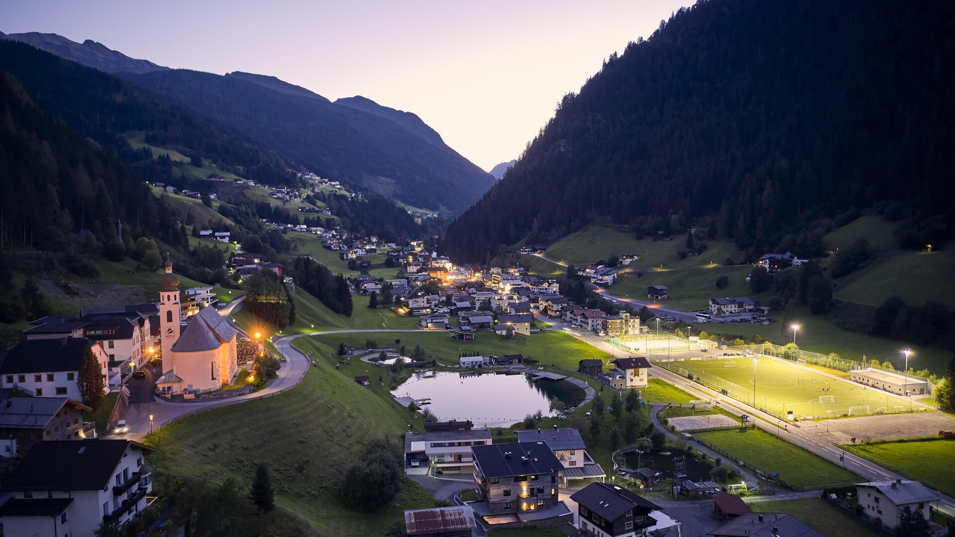Eine malerische Berglandschaft mit einem ruhigen Tal und einem kleinen Dorf. Die Umgebung ist bei Dämmerung beleuchtet und zeigt eine friedliche Atmosphäre.