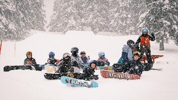 Eine Gruppe von Snowboardern sitzt im Schnee während eines Schneesturms. Um sie herum sind verschneite Bäume und eine winterliche Landschaft.