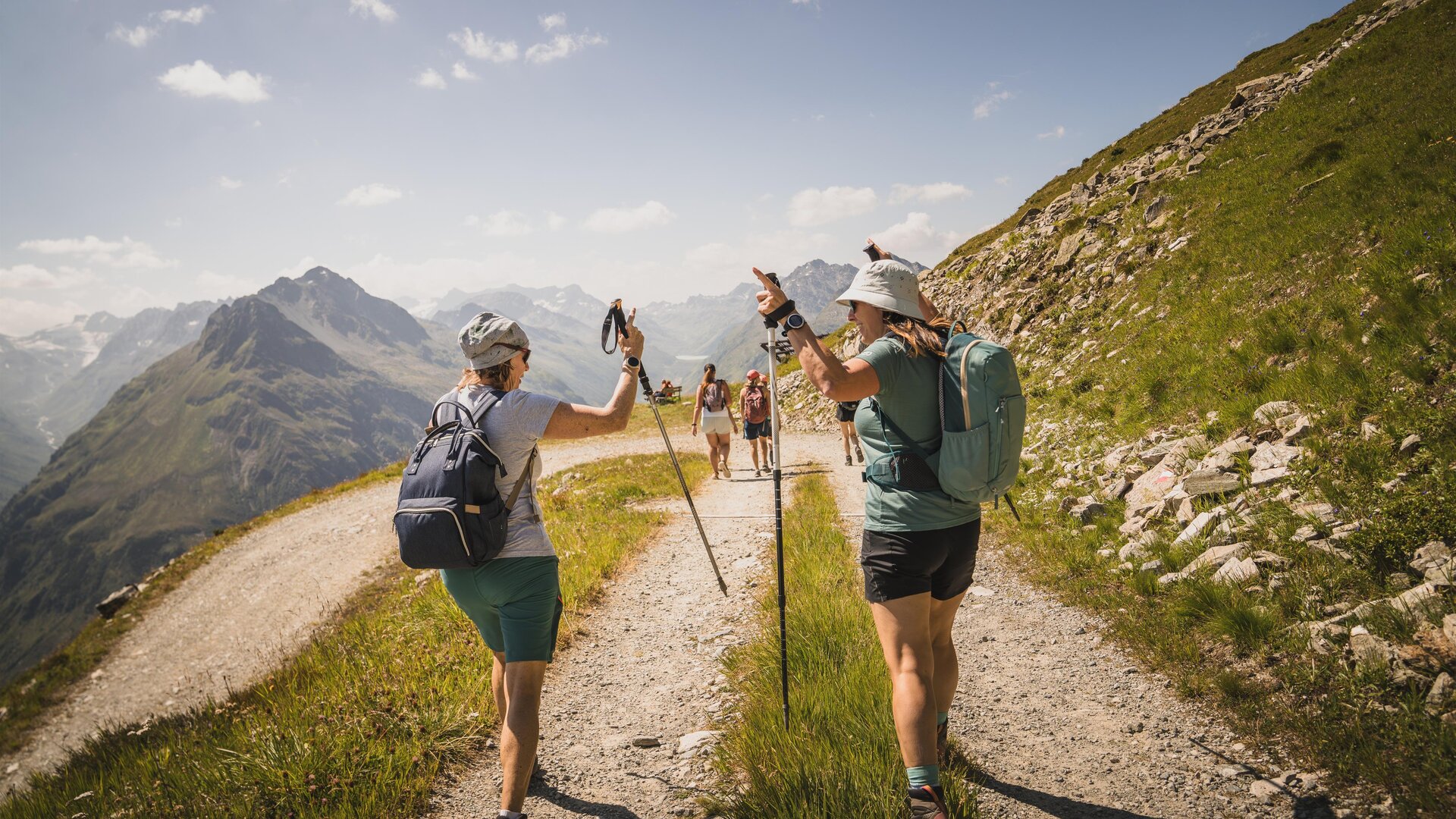Zwei Wanderer auf einem Bergweg genießen die Natur. Im Hintergrund sind weitere Wanderer und Berge zu sehen.
