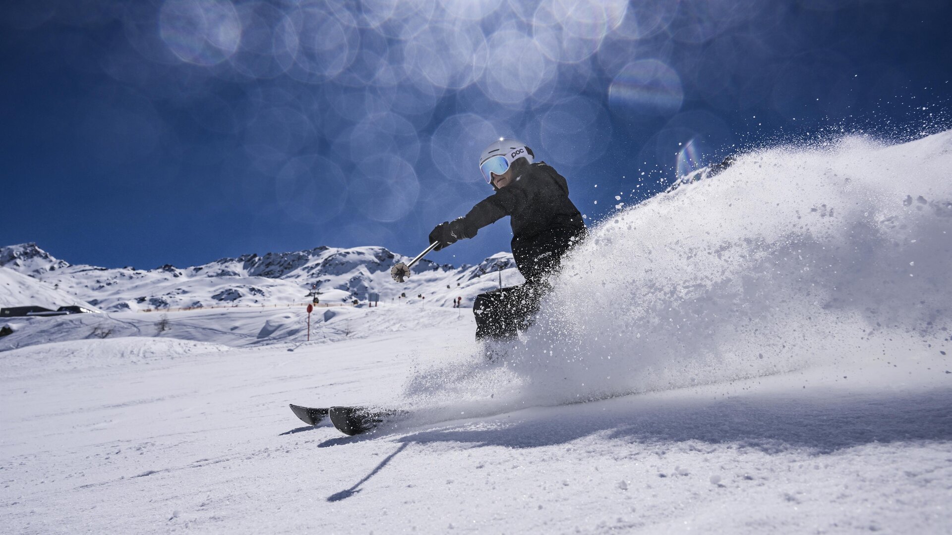 Ein Skifahrer fährt mit Schwung durch den frischen Schnee. Im Hintergrund sind schneebedeckte Berge und ein klarer blauer Himmel zu sehen.