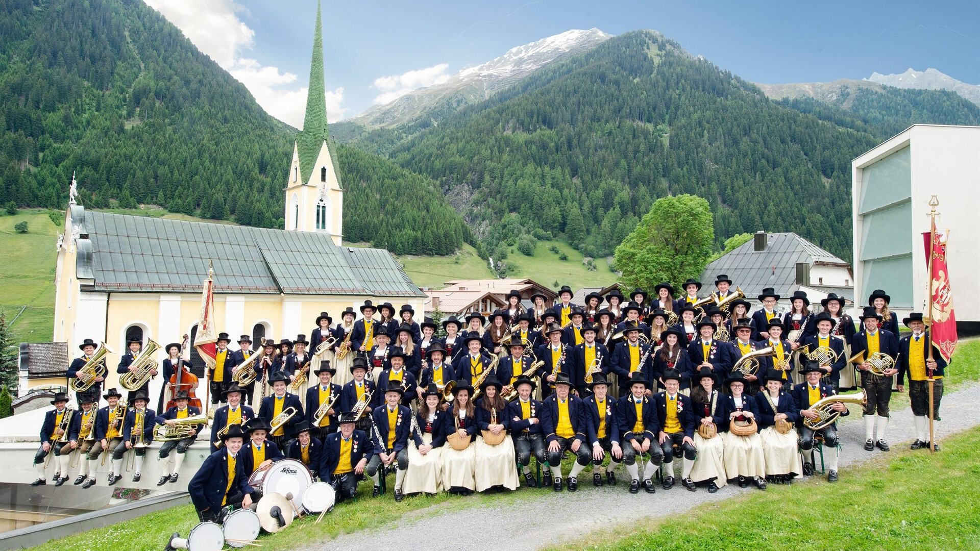Eine große Gruppe von Menschen in traditionellen Trachten steht in einer malerischen Landschaft. Im Hintergrund sieht man eine Kirche und die Berge der Alpen.