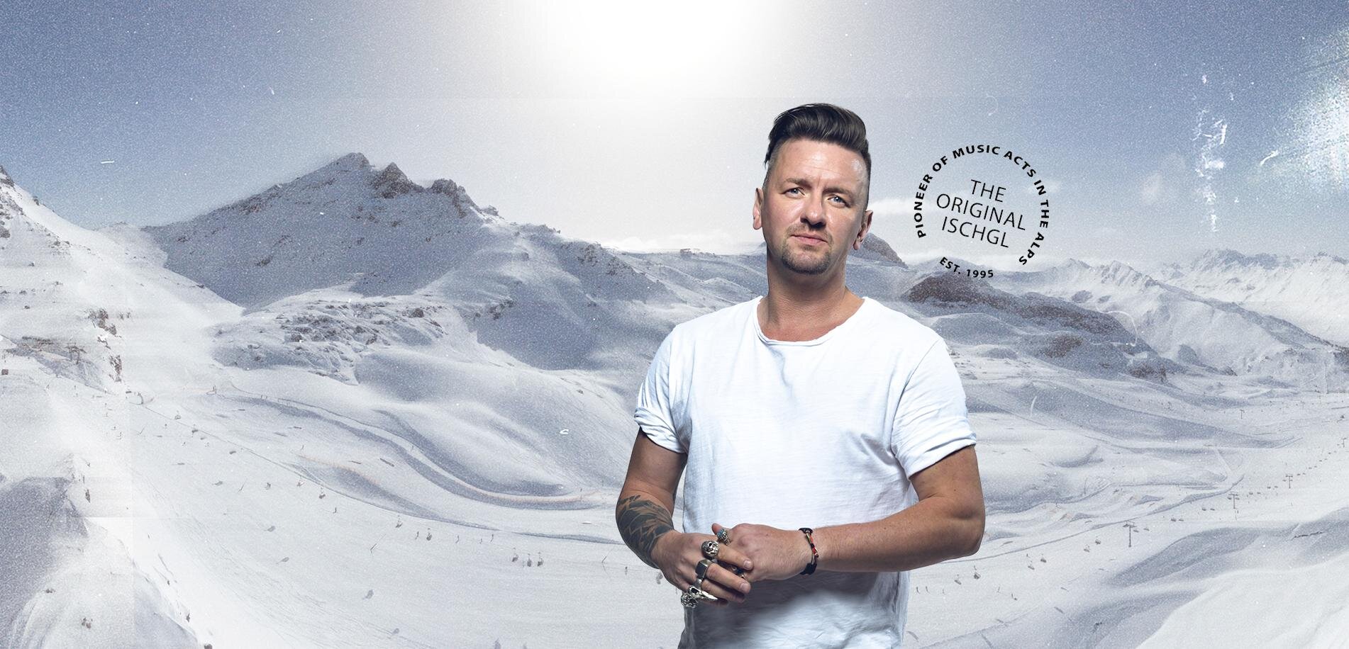 Ein Mann steht in einer schneebedeckten Berglandschaft. Die Umgebung ist weiß und frostig mit hohen Gipfeln im Hintergrund.