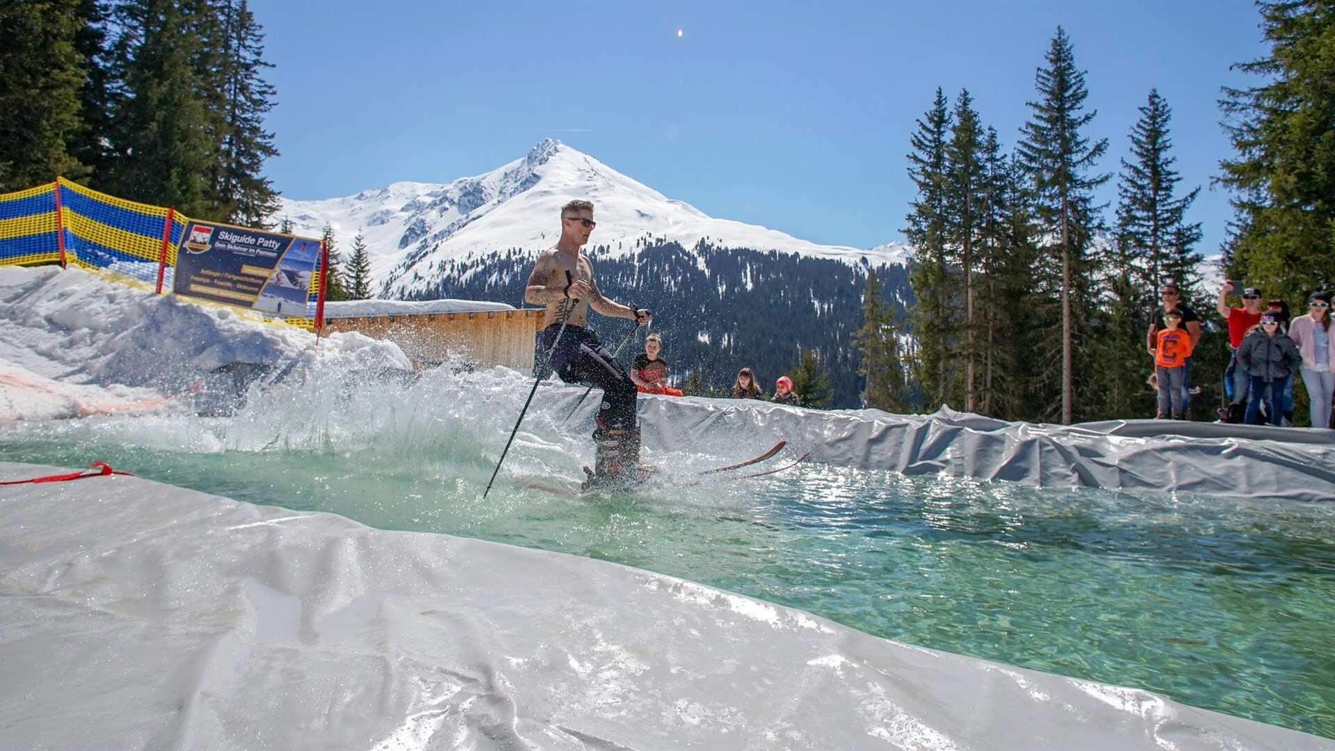 Ein Skifahrer fährt über einen Wassersprung in einer alpinen Umgebung. Im Hintergrund sind schneebedeckte Berge und Tannenbäume zu sehen.