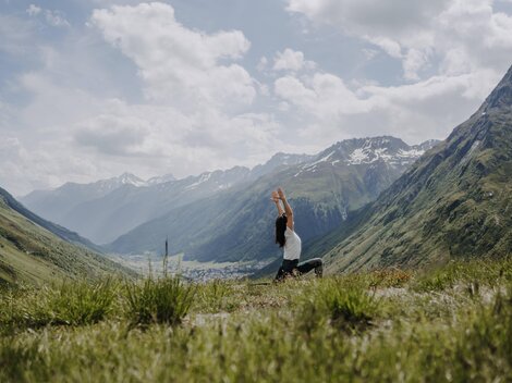 A person is practicing yoga in a picturesque mountain landscape. The surroundings are green and the mountains are partially snow-covered under a clear sky.