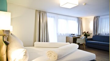 Ein modernes Hotelzimmer mit einem großen Bett, frischen Handtüchern und einem gemütlichen Sofa. Die Fenster lassen viel Tageslicht herein und sorgen für eine einladende Atmosphäre.