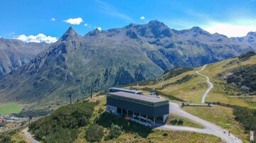 Ein modernes Gebäude in den Bergen, umgeben von grünen Wiesen und hohen Gipfeln. Der Himmel ist klar und blau, ideal für Outdoor-Aktivitäten.