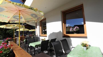 Eine einladende Terrasse mit Tischen und Stühlen. Sonnenschirme spenden Schatten und Blumen schmücken den Raum.