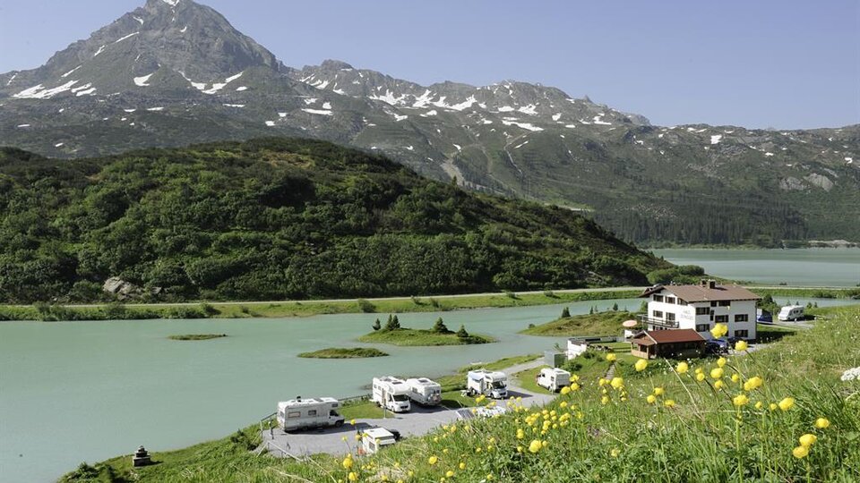 Eine malerische Landschaft mit einem ruhigen See und einem beeindruckenden Berg im Hintergrund. Am Ufer stehen Wohnmobile und ein Gasthaus umgeben von grünen Wiesen.