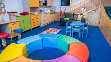 Ein farbenfroher Spielbereich mit einer bunten Sitzgelegenheit in Form eines Kreises. Im Hintergrund gibt es eine Küche und gemütliche Sitzecke für Kinder.