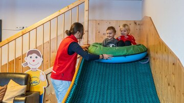 Ein Raum mit einer Rutsche, auf der zwei Kinder sitzen. Eine Betreuerin hilft ihnen beim Rutschen.