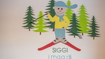 Ein fröhlicher Skifahrer mit einer blauen Mütze und gelbem Pullover steht auf roten Skiern. Im Hintergrund sind grüne Bäume zu sehen.