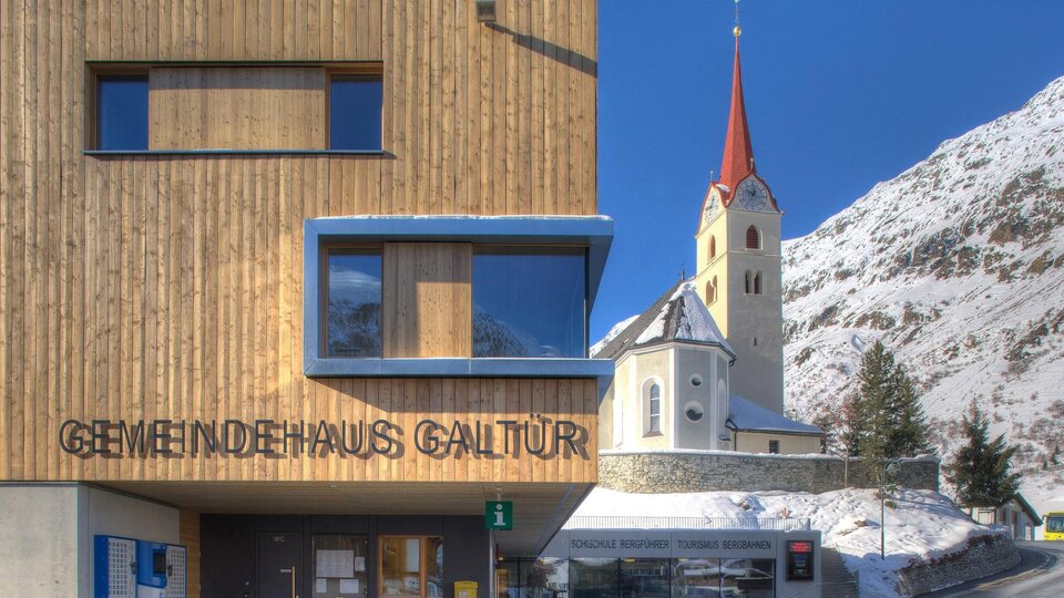 Ein modernes Gemeindehaus mit Holzfassade in Galtür. Im Hintergrund ist eine Kirche mit rotem Kirchturm und schneebedeckte Berge zu sehen.