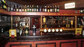 Eine gemütliche Bar mit einer langen Theke und vielen Getränken. Über der Theke hängen verschiedene Flaschen und ein Schild mit einer freundlichen Botschaft.