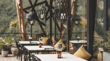 Ein gemütlicher Außenbereich eines Restaurants mit Holzdecks und modernen Möbeln. Die Tische sind für Gäste vorbereitet und es gibt dekorative Pflanzen.