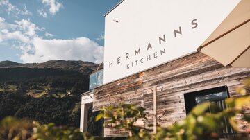 Eine moderne Restaurantfassade mit dem Schriftzug "Hermann's Kitchen". Im Hintergrund sind Berge und ein blauer Himmel zu sehen.