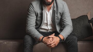 Ein lächelnder Mann sitzt auf einer Couch vor einer Wand mit Weinen. Er trägt einen grauen Blazer und weiße Sneakers.