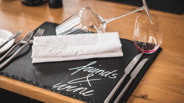 Ein stilvoll gedeckter Tisch mit einem Weinglas und einem umgekippten Glased. Auf dem Tisch liegt eine Serviette und eine Schwarzplatte mit der Aufschrift "Friends & Wine".