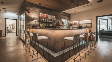 Eine elegante Bar mit einem Holzdesign und stilvollen Hochstühlen. Im Hintergrund sind Regale mit verschiedenen Spirituosen zu sehen.