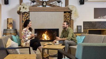 Ein gemütlicher Raum mit einem Kamin und zwei Personen, die sich unterhalten. Sie genießen heiße Getränke und eine entspannte Atmosphäre.