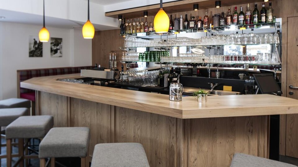 Eine moderne Bar mit einem geschwungenen Holztresen und stilvollen Hockern. Im Hintergrund sind Regale mit einer Auswahl an Spirituosen und Gläsern zu sehen.