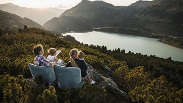 Eine Familie sitzt auf Felsen und schaut auf einen schönen See. Im Hintergrund sind Berge und ein klarer Himmel zu sehen.