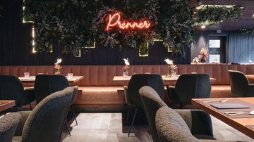 Ein modernes Restaurant mit stilvollen Sitzbereichen und einer einladenden Atmosphäre. Am Hintergrund leuchtet ein Neon-Schriftzug „Prenner“ vor einer grünen Pflanzendekoration.