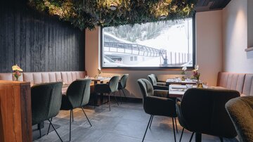 Ein modernes Restaurant mit eleganten, grünen Stühlen und einer gemütlichen Atmosphäre. Durch das große Fenster sieht man eine verschneite Landschaft.