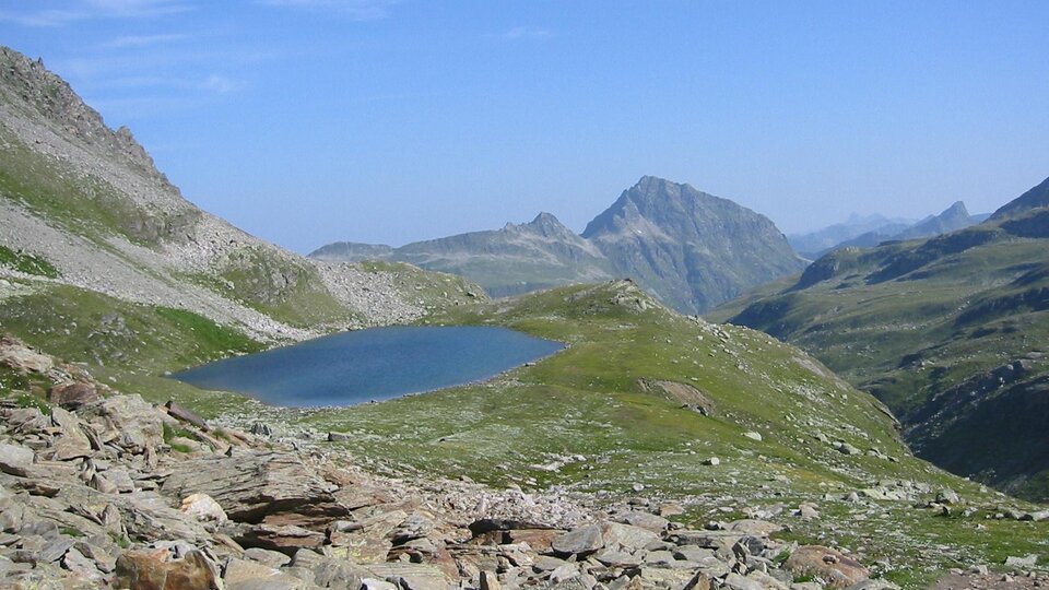 Eine malerische Berglandschaft mit einem klaren, blauen See. Im Hintergrund erheben sich majestätische Berge unter einem blauen Himmel.