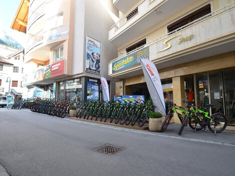 A street with several bicycles available for rent. In the background, shops and advertising flags are visible.