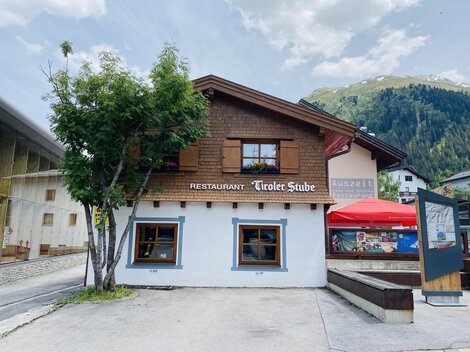 A cozy restaurant called "Tiroler Stube" with wooden paneling and windows. The mountains are visible in the background.
