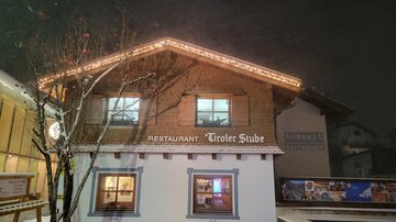 Ein gemütliches Restaurant mit dem Namen "Tiroler Stube", beleuchtet von Lichtern, steht im Schnee. Die Szene vermittelt eine winterliche Atmosphäre bei Nacht.
