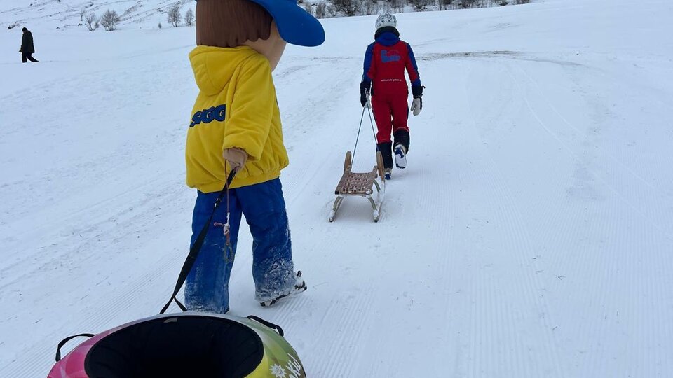 A child is pulling a tube tire up a snowy slope. In the background, you can see other people riding sleds.