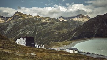Eine malerische Berglandschaft mit einem klaren See im Vordergrund. Im Hintergrund erheben sich beeindruckende Gipfel und Wolken am Himmel.