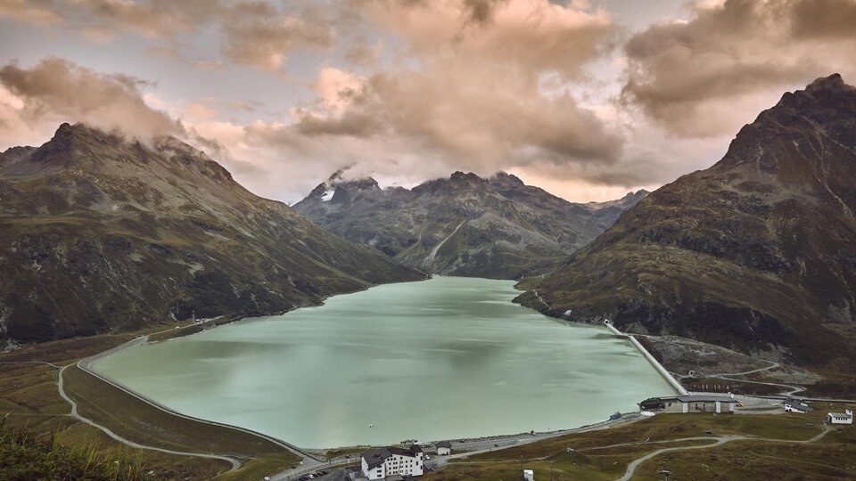 Ein schöner Bergsee, umgeben von hohen Bergen und dramatischen Wolken. Die ruhige Wasseroberfläche spiegelt die umliegende Landschaft wider.