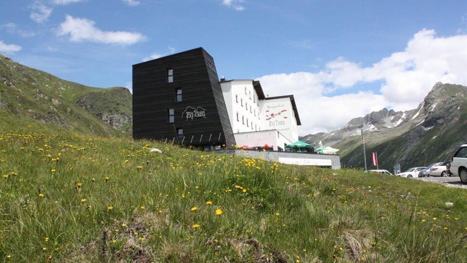Ein modernes Gebäude in den Bergen, umgeben von grünen Wiesen und blühenden Blumen. Im Hintergrund sind majestätische Berge und ein blauer Himmel zu sehen.