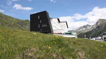Ein modernes Gebäude in den Bergen, umgeben von grünen Wiesen und blühenden Blumen. Im Hintergrund sind majestätische Berge und ein blauer Himmel zu sehen.