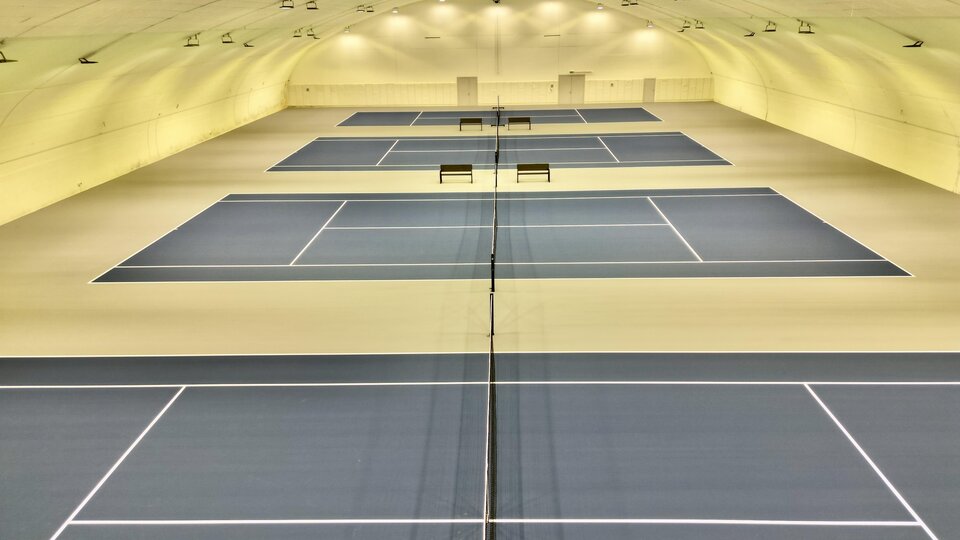 A modern tennis hall with several playing fields. The blue courts are well-lit and provide space for multiple games.